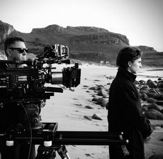 a man standing next to a woman on top of a beach near a camera set