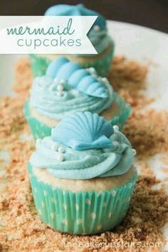 three cupcakes with blue frosting sitting on top of a white plate covered in sand