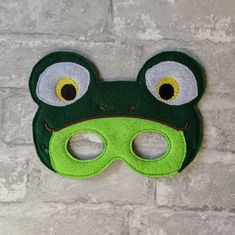 a green frog mask with yellow eyes on a white brick wall behind it is a hole in the felt