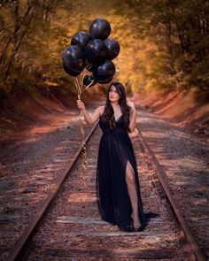 a woman in a long black dress holding balloons on train tracks with trees behind her