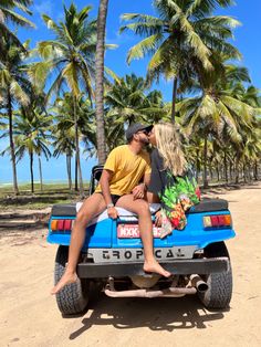 a man and woman sitting in the back of a blue jeep on a sandy beach