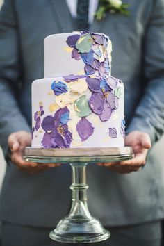 a close up of a person holding a cake on a metal stand with purple flowers