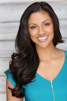 a woman with long dark hair wearing a blue shirt and smiling at the camera while standing in front of a wooden wall