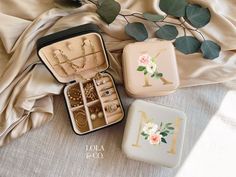 three small jewelry cases sitting on top of a white cloth next to some leaves and flowers