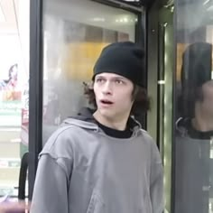 a young man wearing a beanie standing in front of a glass door with his mouth open