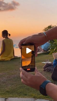 two people sitting on the grass and one person holding up a cell phone