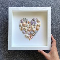 a hand holding a white frame with sea shells in the shape of a heart on it