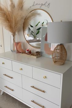 a white dresser topped with lots of drawers next to a mirror and vase filled with feathers