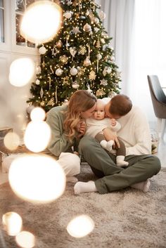 a man and woman sitting on the floor with a baby in front of a christmas tree