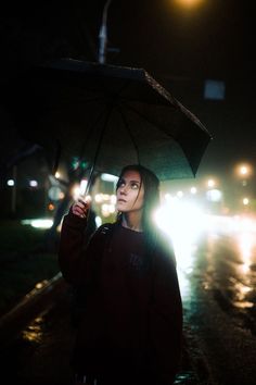 a woman holding an umbrella on the side of a road at night with street lights in the background