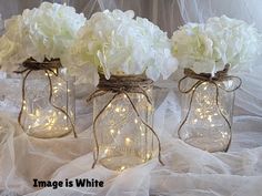 three mason jars filled with white flowers and string lights are sitting on a lace covered tablecloth