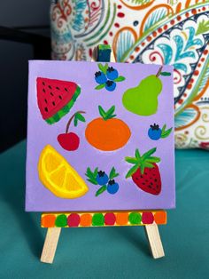 an easel with fruit painted on it sitting on a blue surface next to pillows