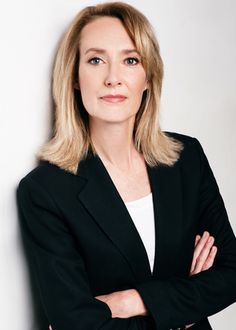 a woman with her arms crossed standing in front of a white wall wearing a black jacket