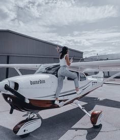 a woman standing on the wing of a small plane in front of an airplane hangar