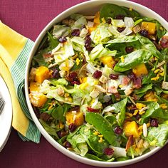 a white bowl filled with salad next to a yellow napkin and silver fork on top of a purple table cloth