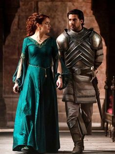 a man and woman dressed in medieval costumes walking together on the street, with one wearing a green dress