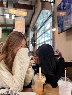 two women sitting at a table with drinks in front of them and one holding her face to her mouth