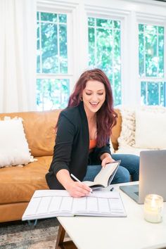 a woman is sitting on the couch and working on her laptop while holding a book