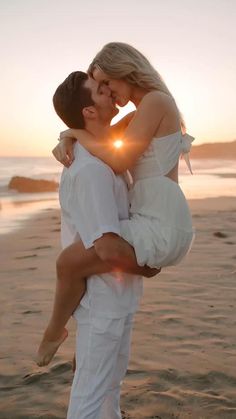 a man and woman kissing on the beach at sunset with the sun setting in the background