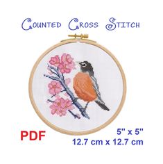 a cross stitch bird sitting on top of a branch with pink flowers in front of it