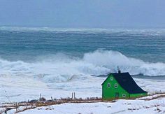 a green house sitting on top of snow covered ground next to the ocean with crashing waves