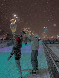 two people are skating on an ice rink in the city at night with snow falling all around them