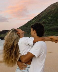 a man holding a woman on the beach at sunset with mountains in the background and pink sky