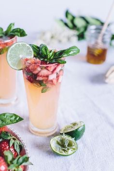 two glasses filled with drinks and garnishes on top of a white table