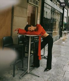 a woman sitting at a table in front of a cafe wearing an orange shirt and black pants