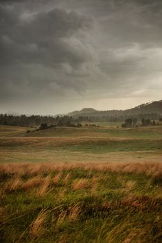 an open field with grass and hills in the background under a cloudy sky on a stormy day