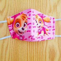 a pink paper face mask with paw patrol characters on the front and back sides, hanging from a wooden table