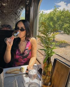 a woman sitting at a table drinking from a wine glass while looking out the window