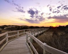 a wooden walkway leading to the beach at sunset with clouds in the sky above it