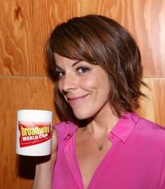 a woman holding a cup in her right hand and smiling at the camera while standing next to a wooden wall