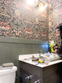 a woman is painting the walls in a bathroom with floral wallpaper and a white toilet