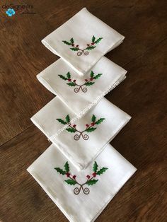 four white christmas napkins with holly and berries on them sitting on top of a wooden table