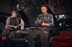 two men are working on an automobile in a garage, one is holding a camera and the other is looking at it