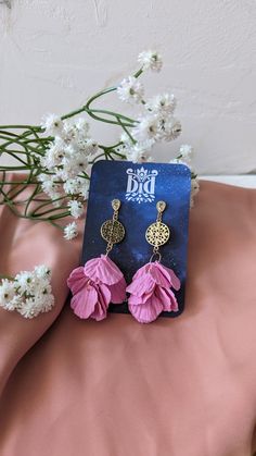 a pair of pink flower earrings sitting on top of a pink sheet next to white flowers