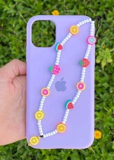 a person holding up a phone case with fruit and beads attached to it in front of some flowers