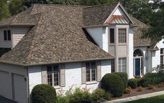 a white house with brown shingles on the roof and bushes in front of it