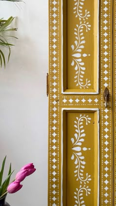 a yellow painted cabinet with white designs on the front and side doors, next to a pink flower