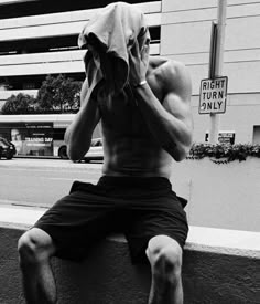 a shirtless man sitting on the side of a building while covering his face with a towel