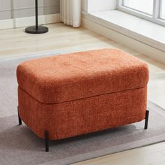 an orange ottoman sitting on top of a rug in front of a window with a lamp next to it