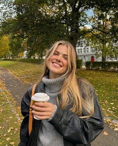 a woman holding a coffee cup in her right hand and walking down a path with trees on both sides