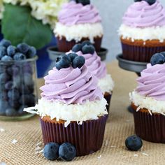 cupcakes with purple frosting and blueberries are on a table next to flowers