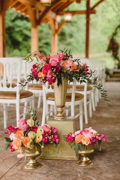 an arrangement of flowers in gold vases on top of a table with white chairs