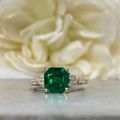 an emerald and diamond ring sits on a table next to a white rose in the background