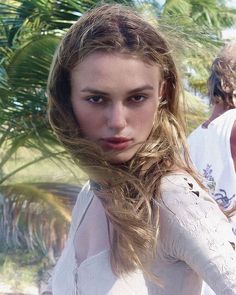 a woman with long hair standing in front of palm trees and looking at the camera