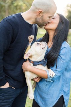 a man and woman kissing while holding a small dog in their lap with trees in the background