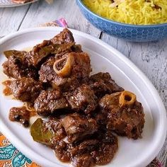 a white plate topped with meat and vegetables next to a bowl filled with yellow rice